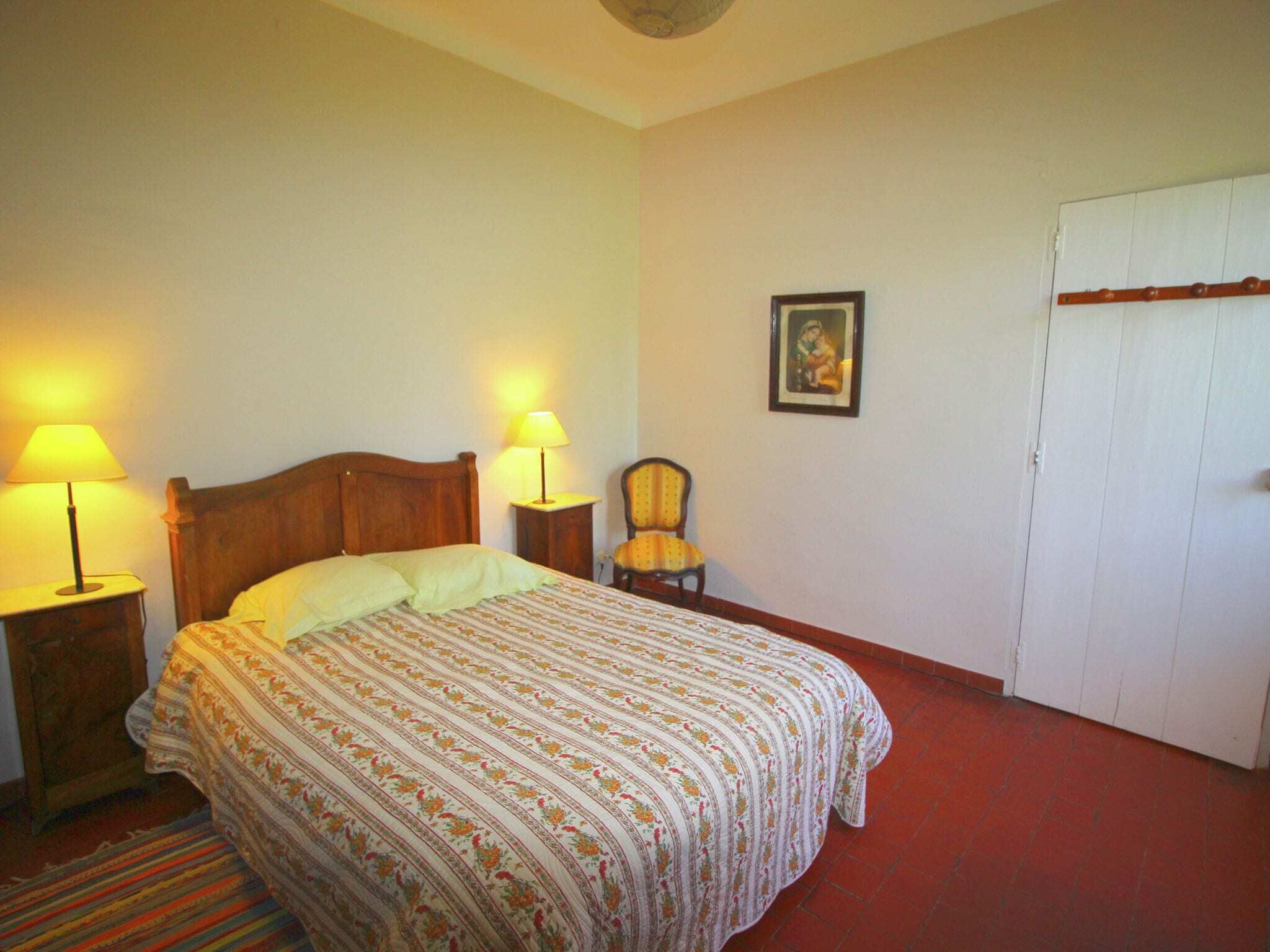 A bedroom with a bed, two bedside lamps, a chair, a door, and a framed picture on the wall.