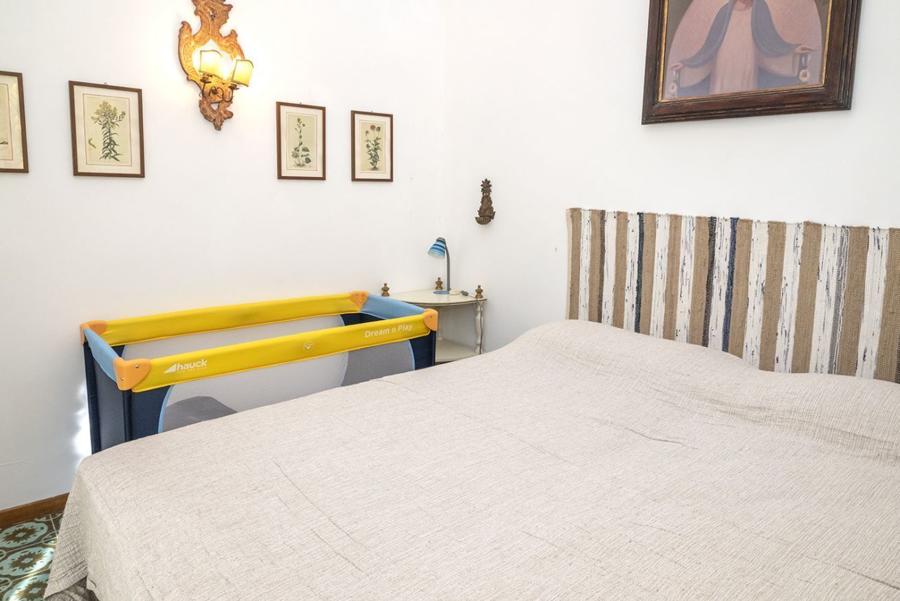A bedroom with a bed, a nightstand, a lamp, a wall-mounted shelf, and framed artwork.