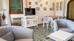 Una sala de estar con un sofá, un televisor sobre un mueble bajo, una mesa de comedor con sillas, un suelo estampado y un armario con vajilla.