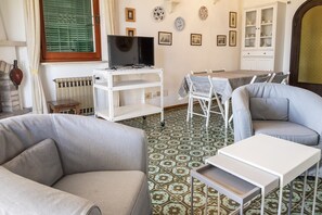 A living room with a sofa, a TV on a stand, a dining table with chairs, a patterned floor, and a cabinet with dishes.