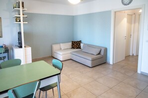 A living room with a sectional sofa, a dining table with chairs, a bookshelf, and a patterned wall.