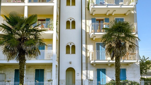 Un edifici de diverses plantes amb balcons, una entrada central i palmeres.