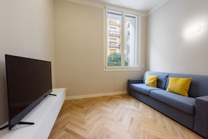 A room with a flat-screen TV on a white stand, a blue sofa with yellow cushions, a window with a view of buildings, and a herringbone-patterned wooden floor.