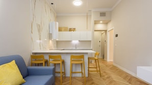 A modern living room with a blue sofa, yellow pillow, and a kitchen area with a white countertop and wooden chairs.