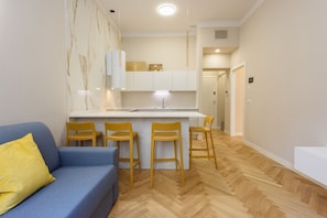 A modern living room with a blue sofa, yellow pillow, and a kitchen area with a white countertop and wooden chairs.