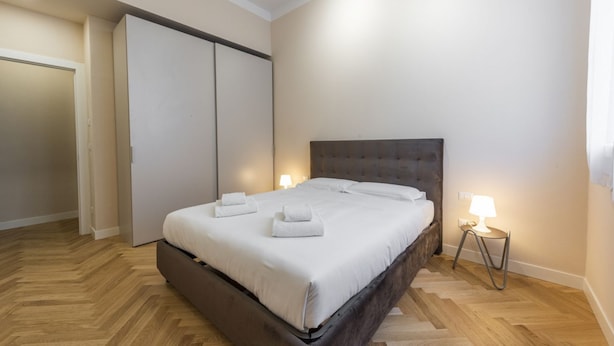 A bedroom with a large bed, a wardrobe, a nightstand with a lamp, and a herringbone-patterned wooden floor.