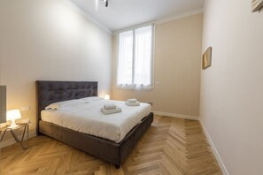 A bedroom with a bed, a window, a lamp, and a herringbone-patterned floor.