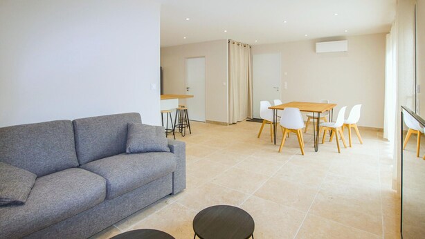 Un salón moderno con un sofá gris, una mesa de centro redonda y un comedor con una mesa de madera y sillas blancas.