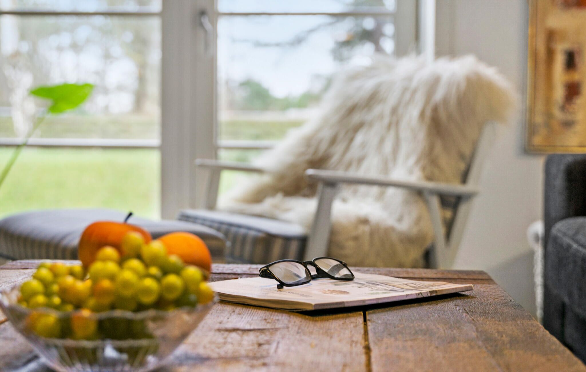 A wooden table with a bowl of fruit, a pair of glasses, and a magazine.
