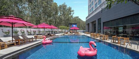 A swimming pool with pink flamingo floats, surrounded by lounge chairs and umbrellas, with a building and trees in the background.