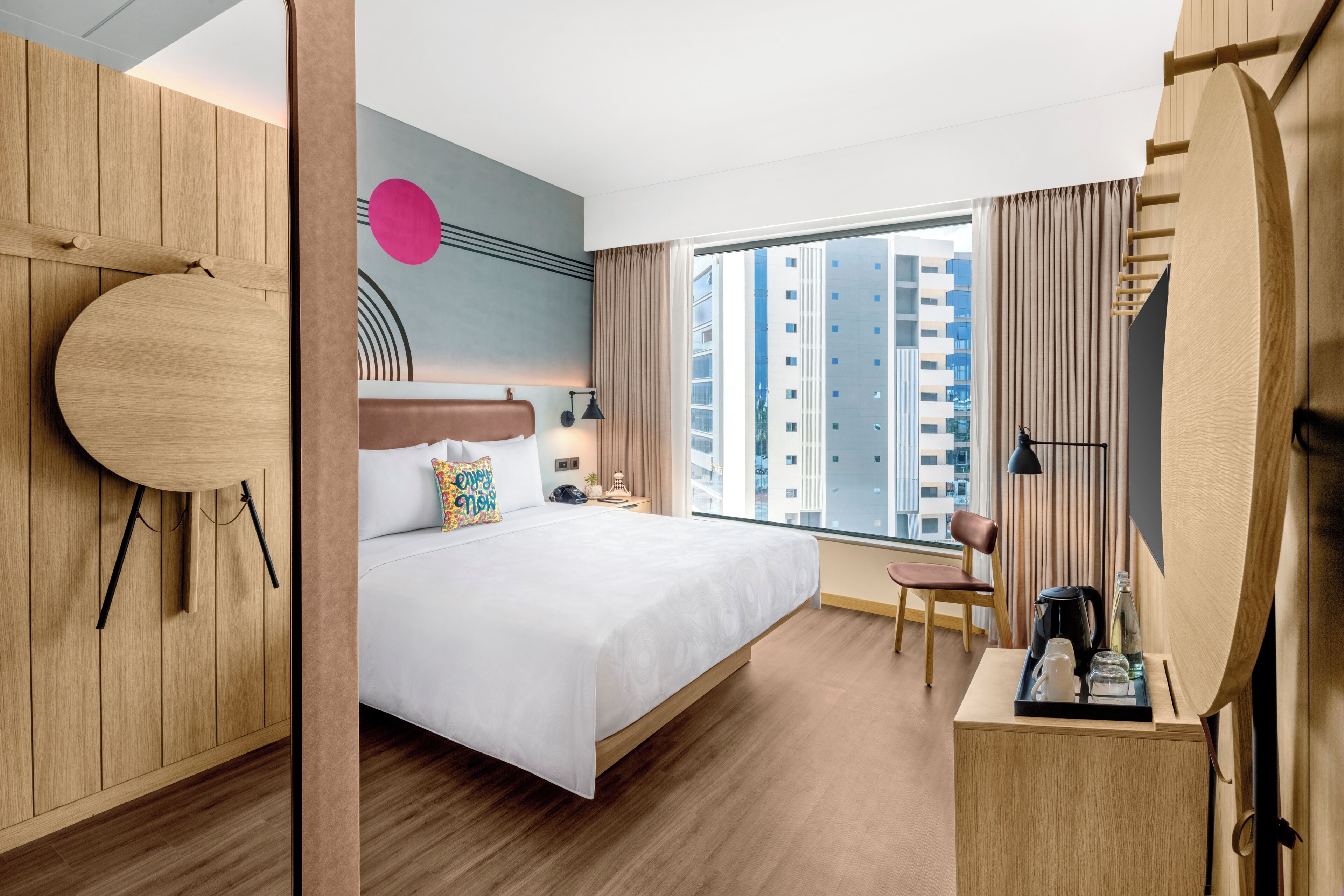 Habitación de hotel con una cama grande, un escritorio con cafetera, una silla, un ventanal con vista a la ciudad y un armario de madera.