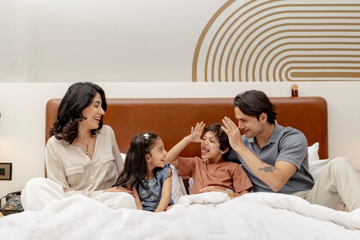 A family of four in a hotel room with a bed, a bedside table, and a wall with a geometric design.