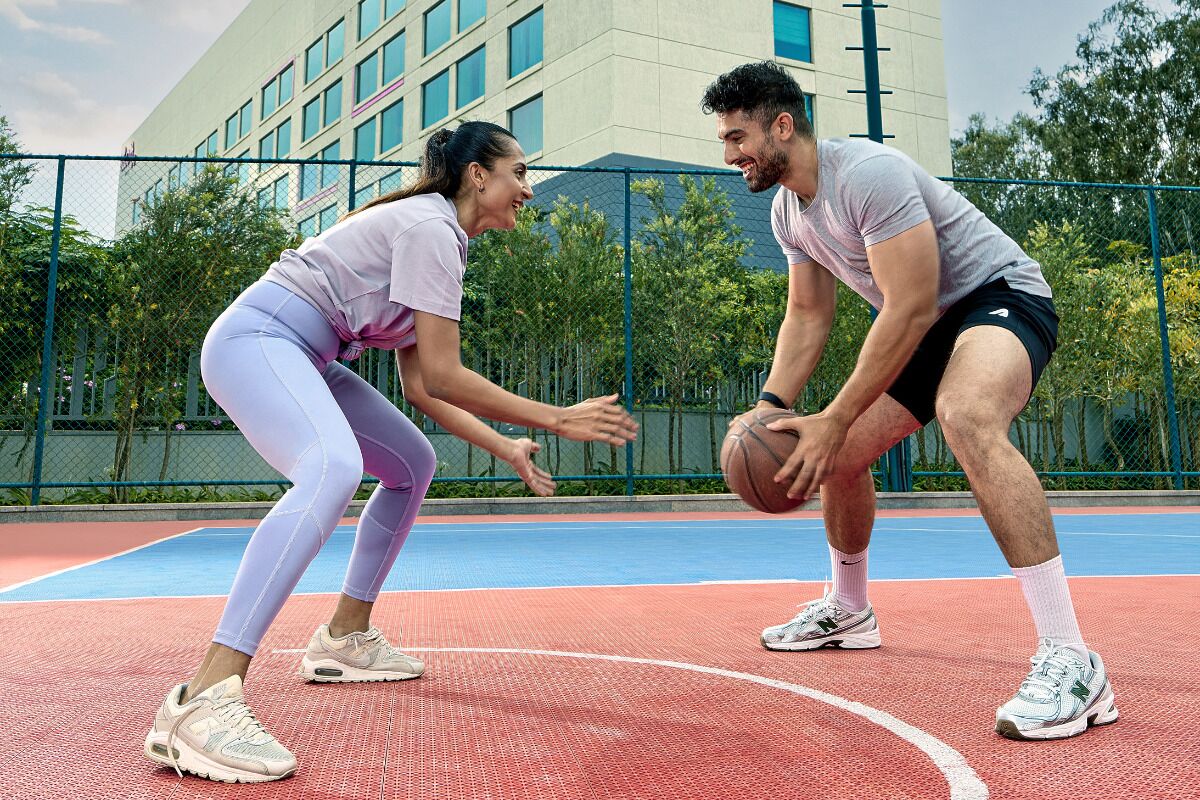 Twee personen spelen basketbal op een buitenbaan, met een gebouw op de achtergrond.