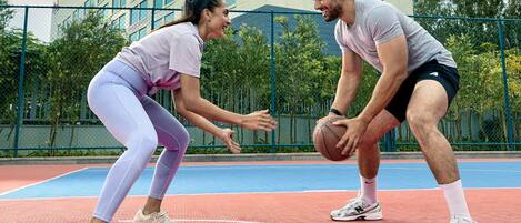 Twee personen spelen basketbal op een buitenbaan, met een gebouw op de achtergrond.