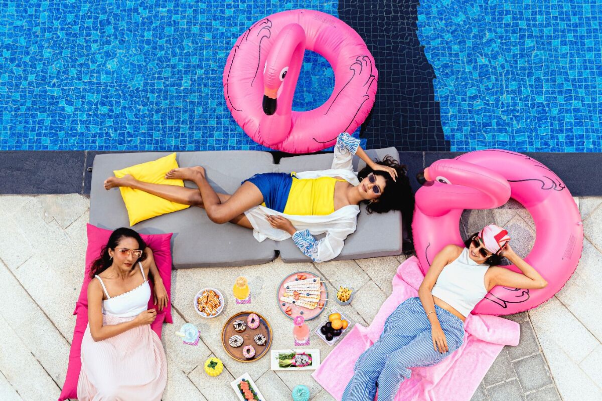 Three people relaxing by a pool with inflatable rings and various food items on the ground.