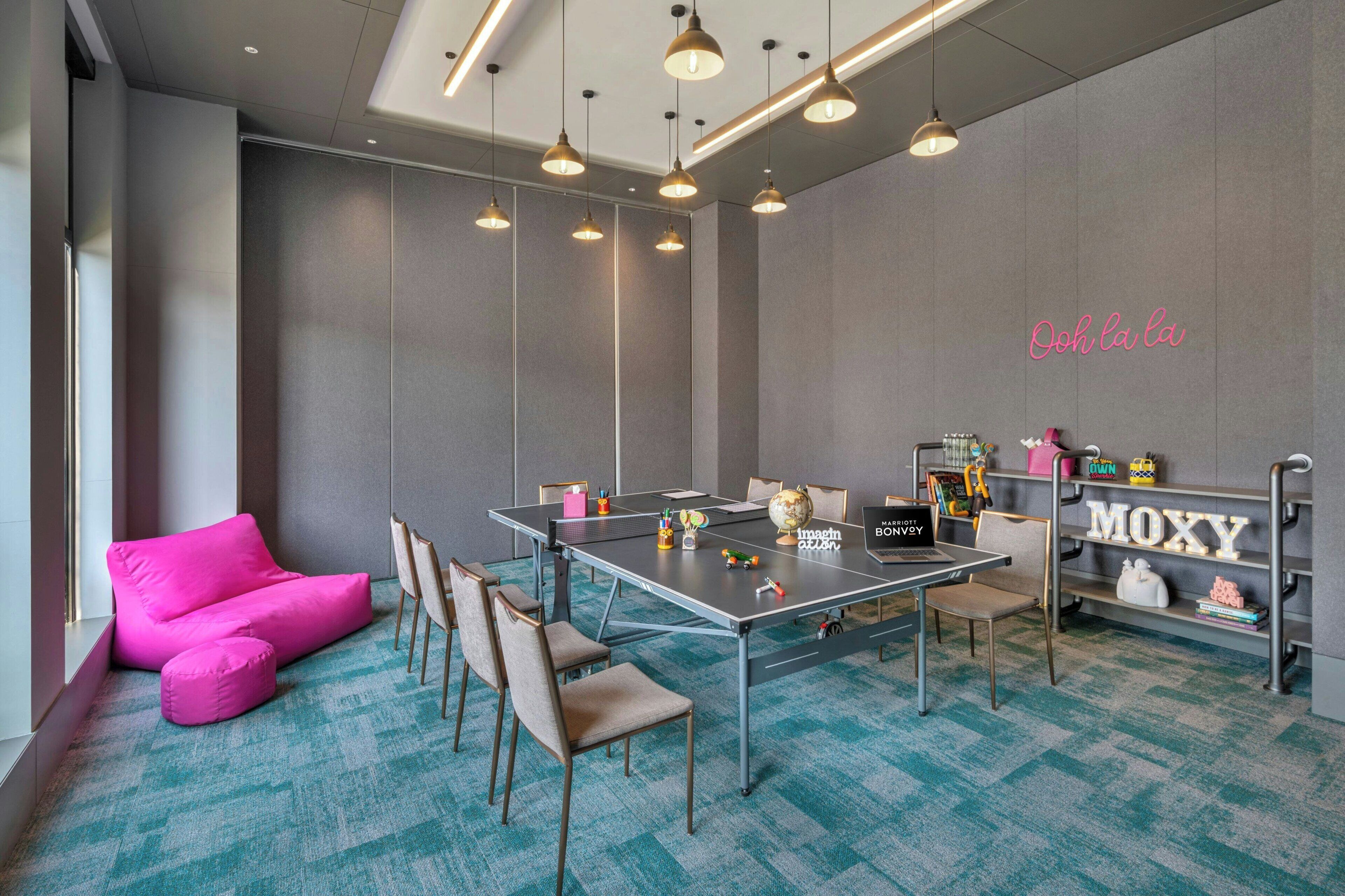 A modern meeting room with a large table, chairs, a pink sofa, and a bookshelf with toys and a globe.