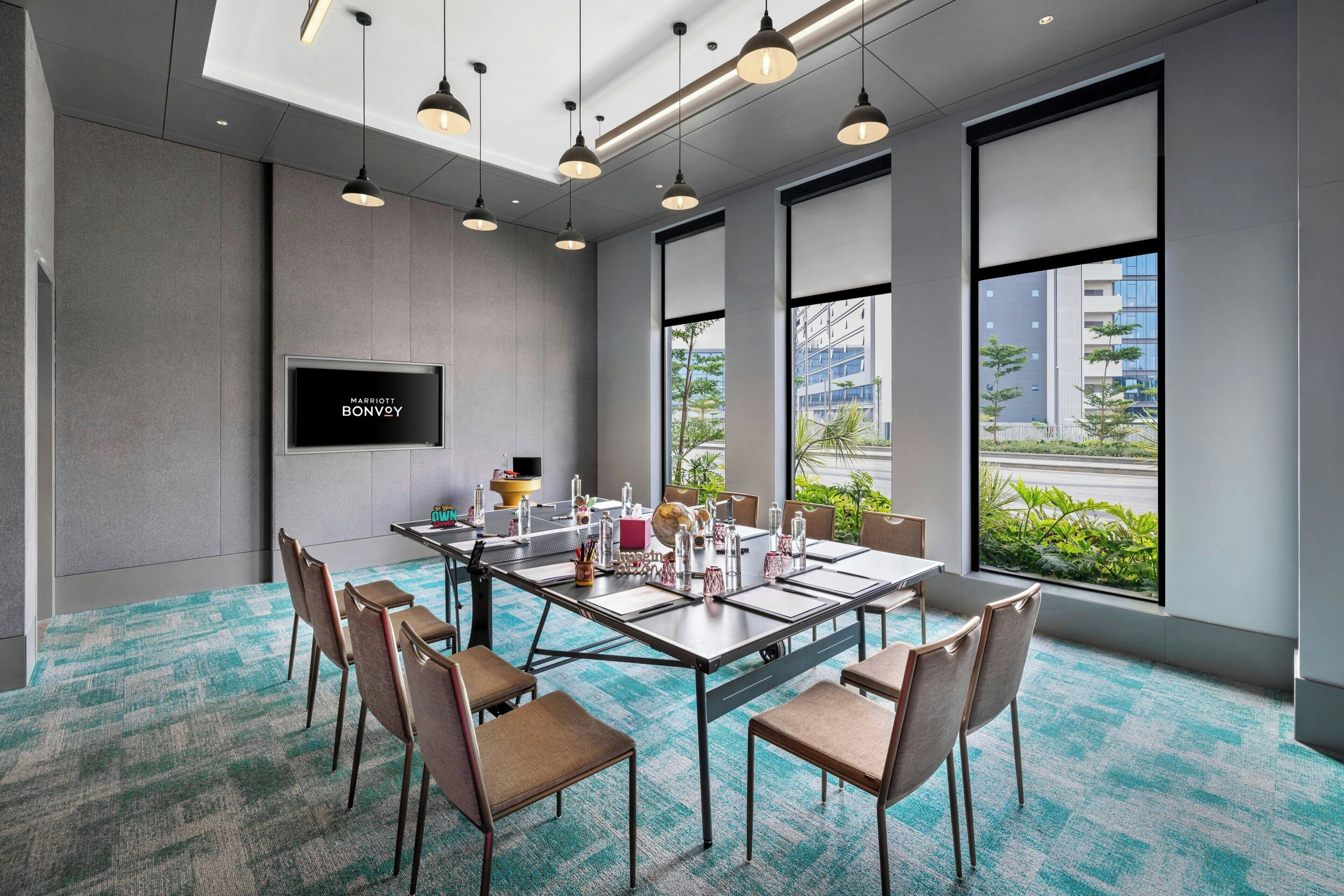 A modern meeting room with a large table, chairs, a flat-screen TV, and large windows with a view of buildings and greenery.