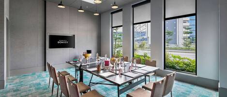 A modern meeting room with a large table, chairs, a flat-screen TV, and large windows with a view of buildings and greenery.
