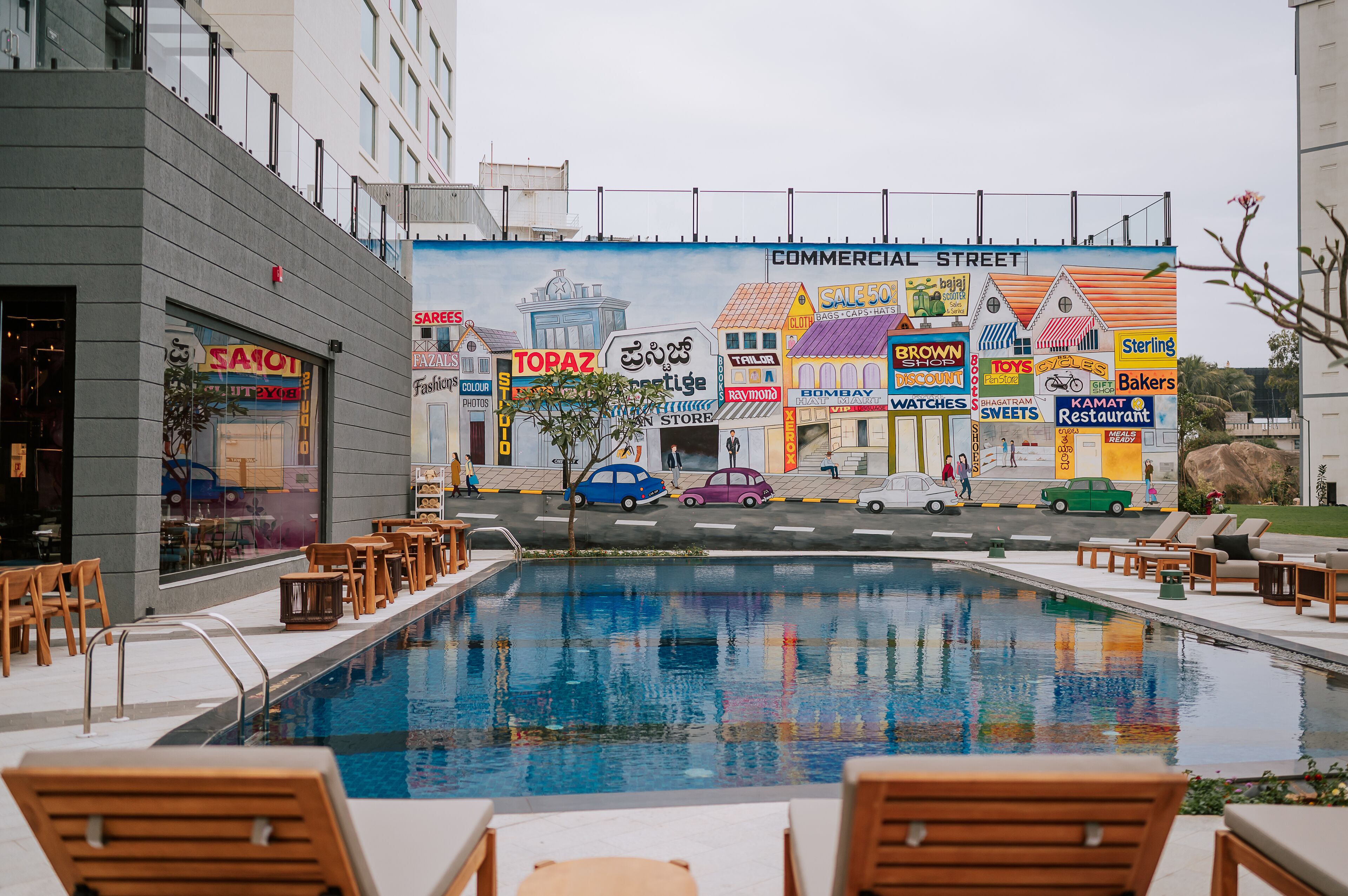 A swimming pool with lounge chairs, a mural of commercial buildings, and a storefront with a sign reading "SAREES".