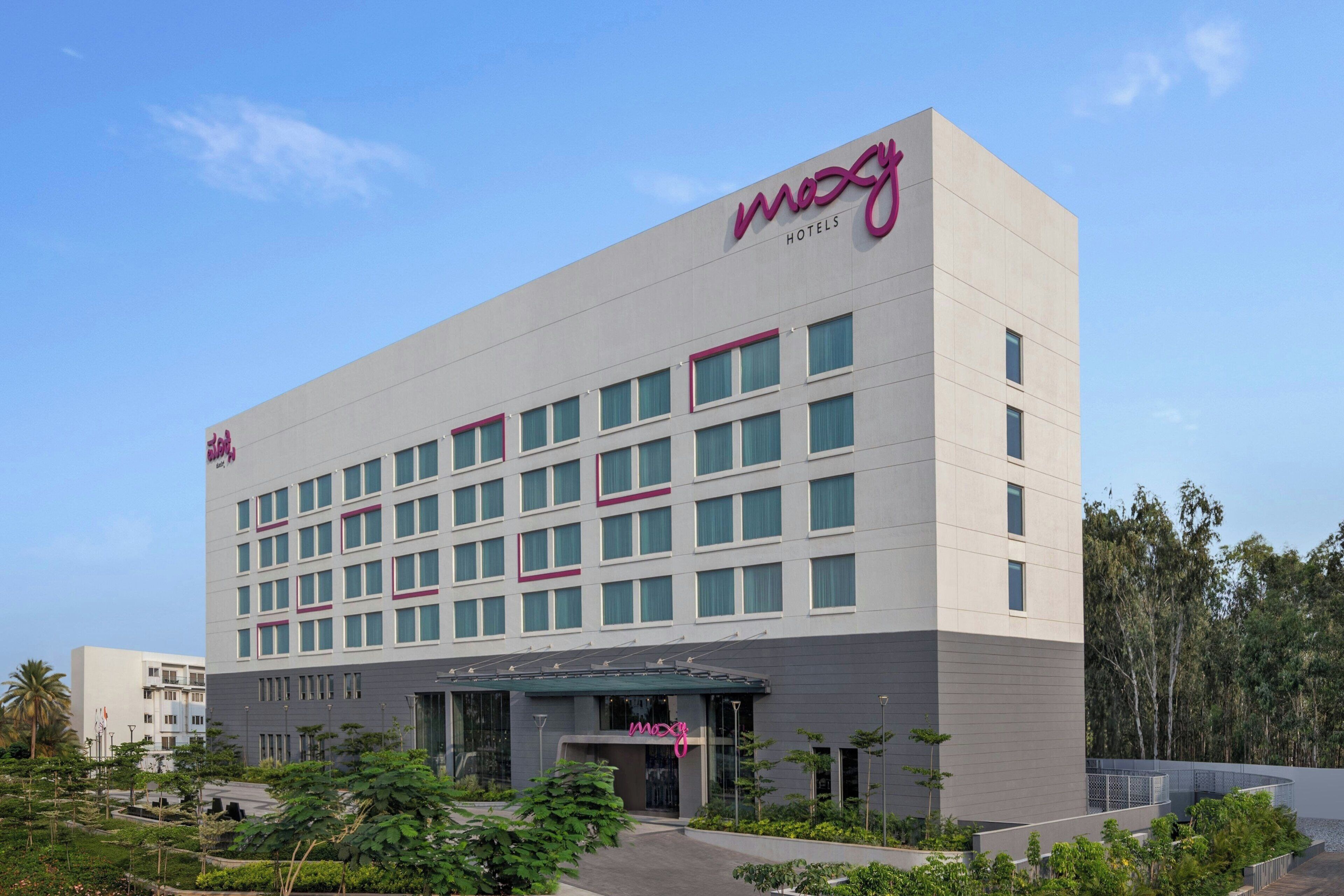 Un moderno edificio de hotel con un llamativo letrero 'mozy', varios pisos y un área de entrada ajardinada.