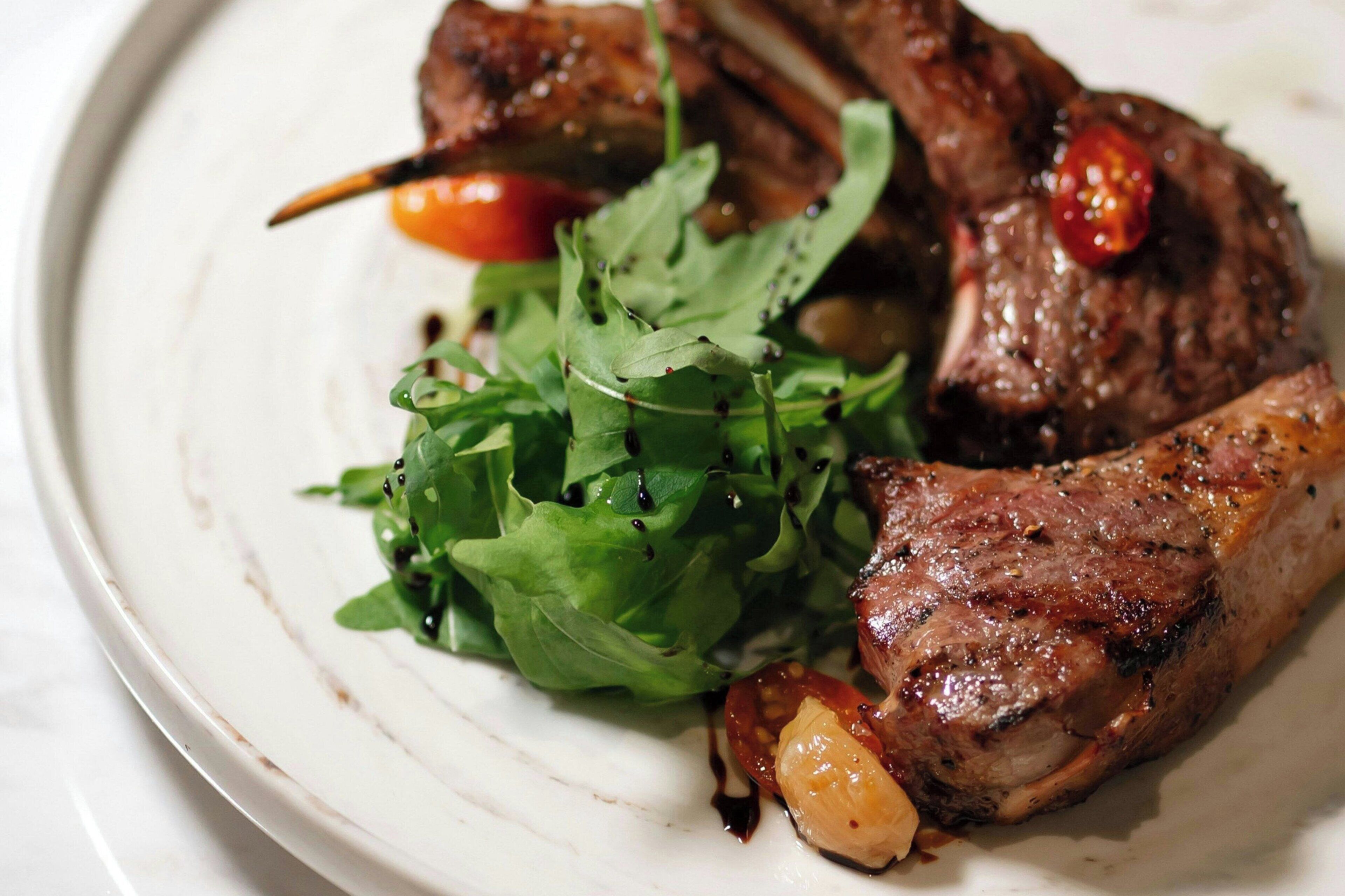 A plate with grilled meat, greens, and a cherry tomato.