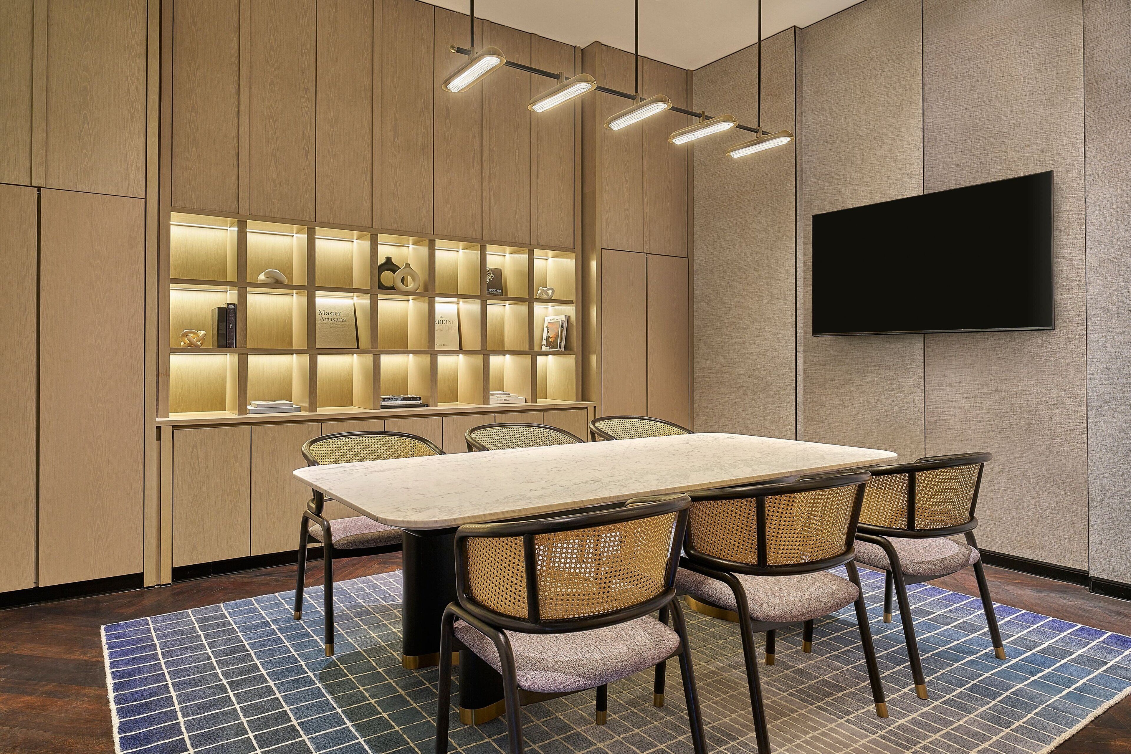A modern conference room with a long table, four wicker chairs, a wall-mounted TV, and illuminated shelves.