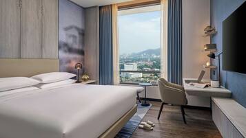 A hotel room with a bed, a desk, a chair, a TV, and a view of the city.