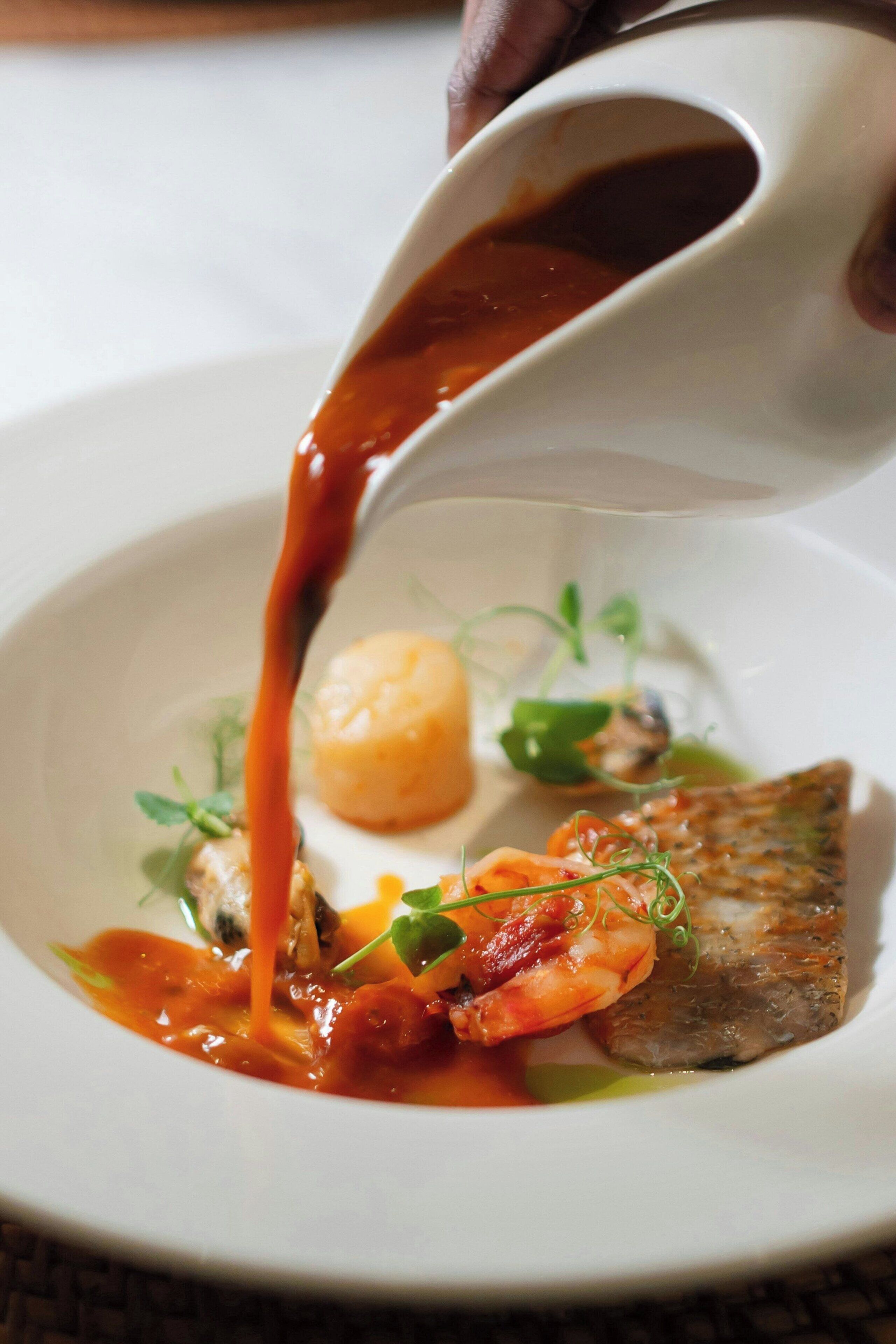 A white plate with a fish, shrimp, and a sauce being poured over it.