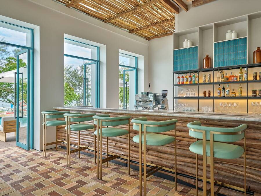 Een bar met houten barstoelen, een toonbank met drankdispenser en planken met flessen en glazen.