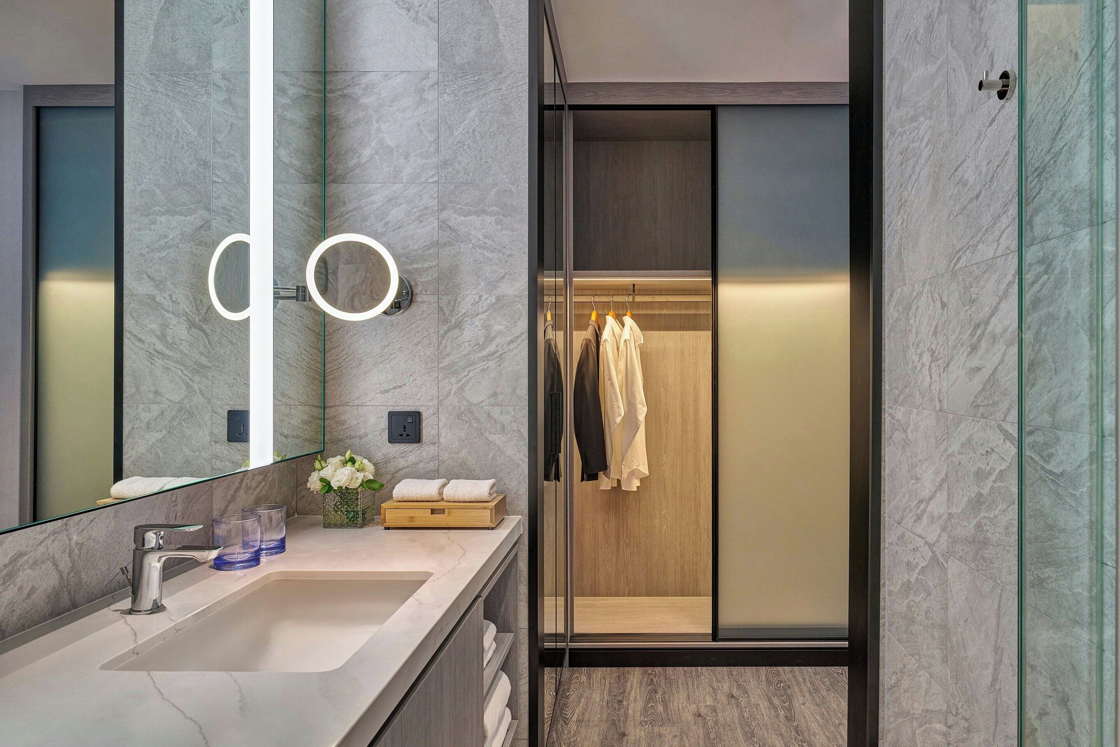 Une salle de bain moderne avec un lavabo en marbre, une douche à parois de verre et un dressing ouvert où les vêtements sont suspendus.