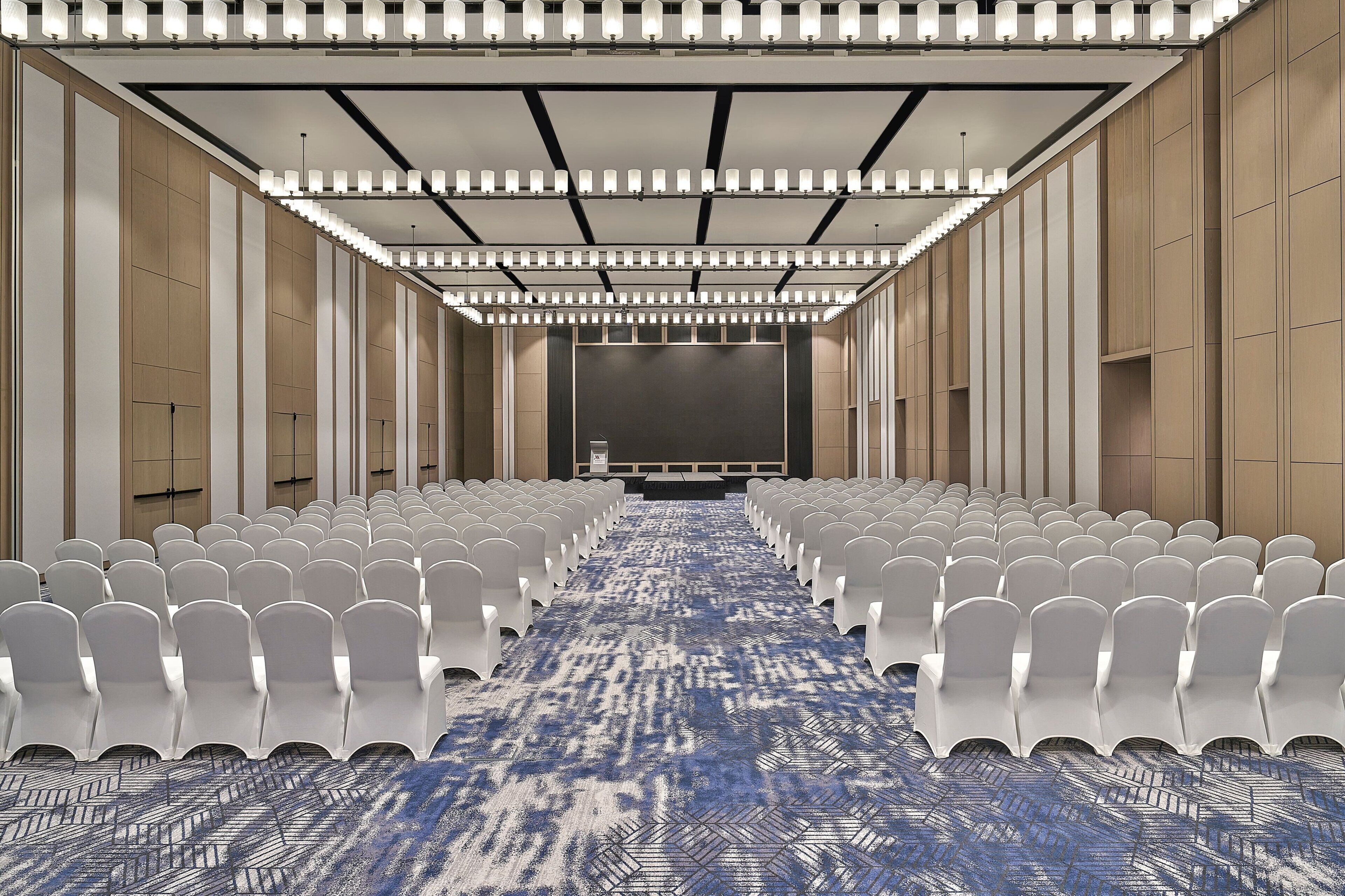 Un salón de conferencias con filas de sillas blancas, una alfombra estampada y una pantalla grande al frente.