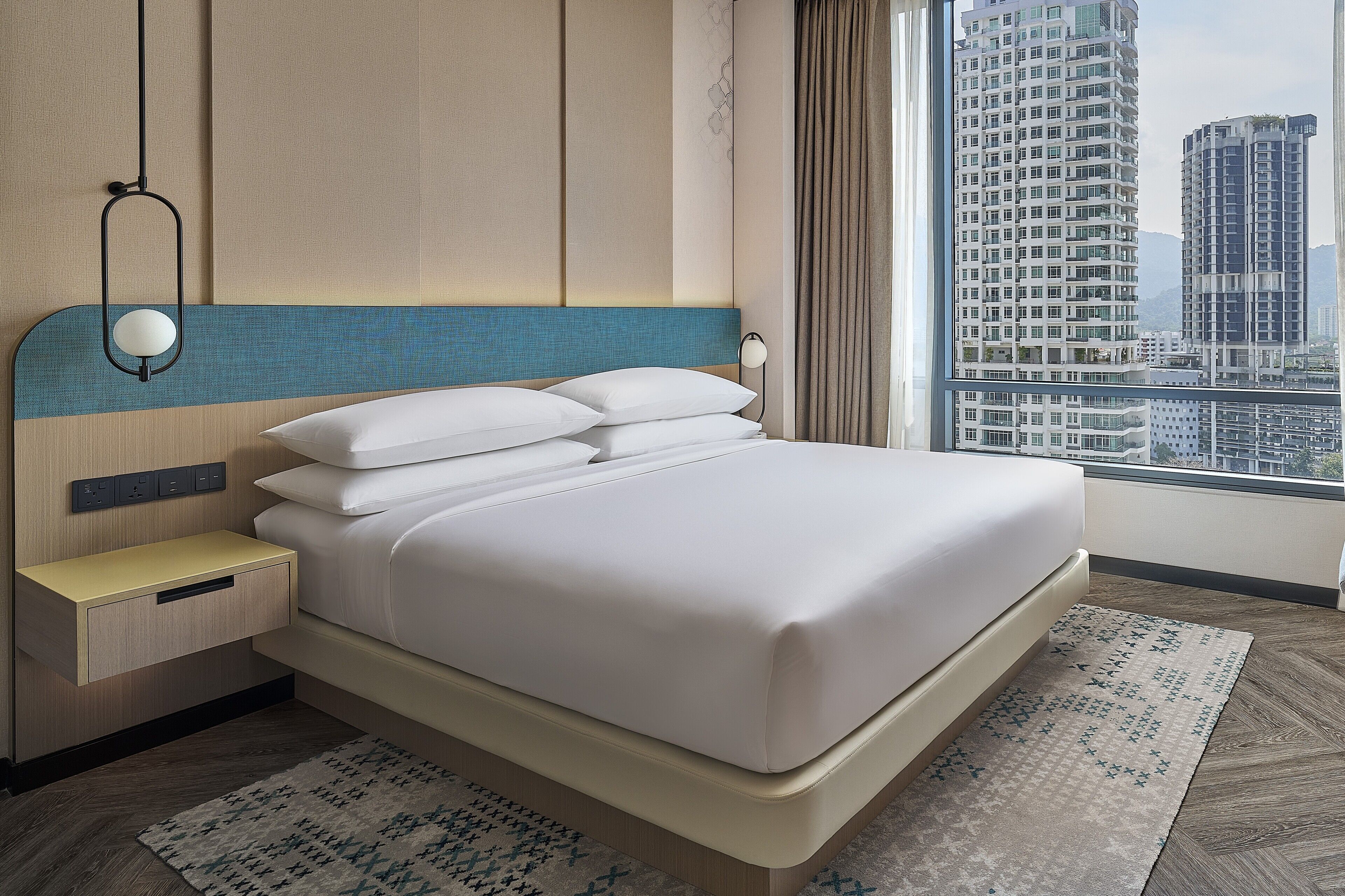 Habitación de hotel moderna con una cama grande, una mesita de noche, una lámpara de pared y vistas de edificios altos a través del ventanal.