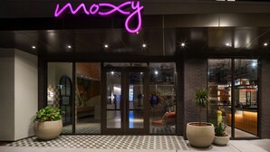 Un edificio moderno con un letrero de neón que dice "moxy" y grandes macetas con plantas a la entrada.