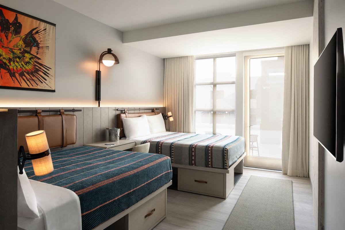 Ein Hotelzimmer mit zwei Betten, einem großen Fenster mit Vorhängen, einem Wand-Fernseher und einem Bild an der Wand.