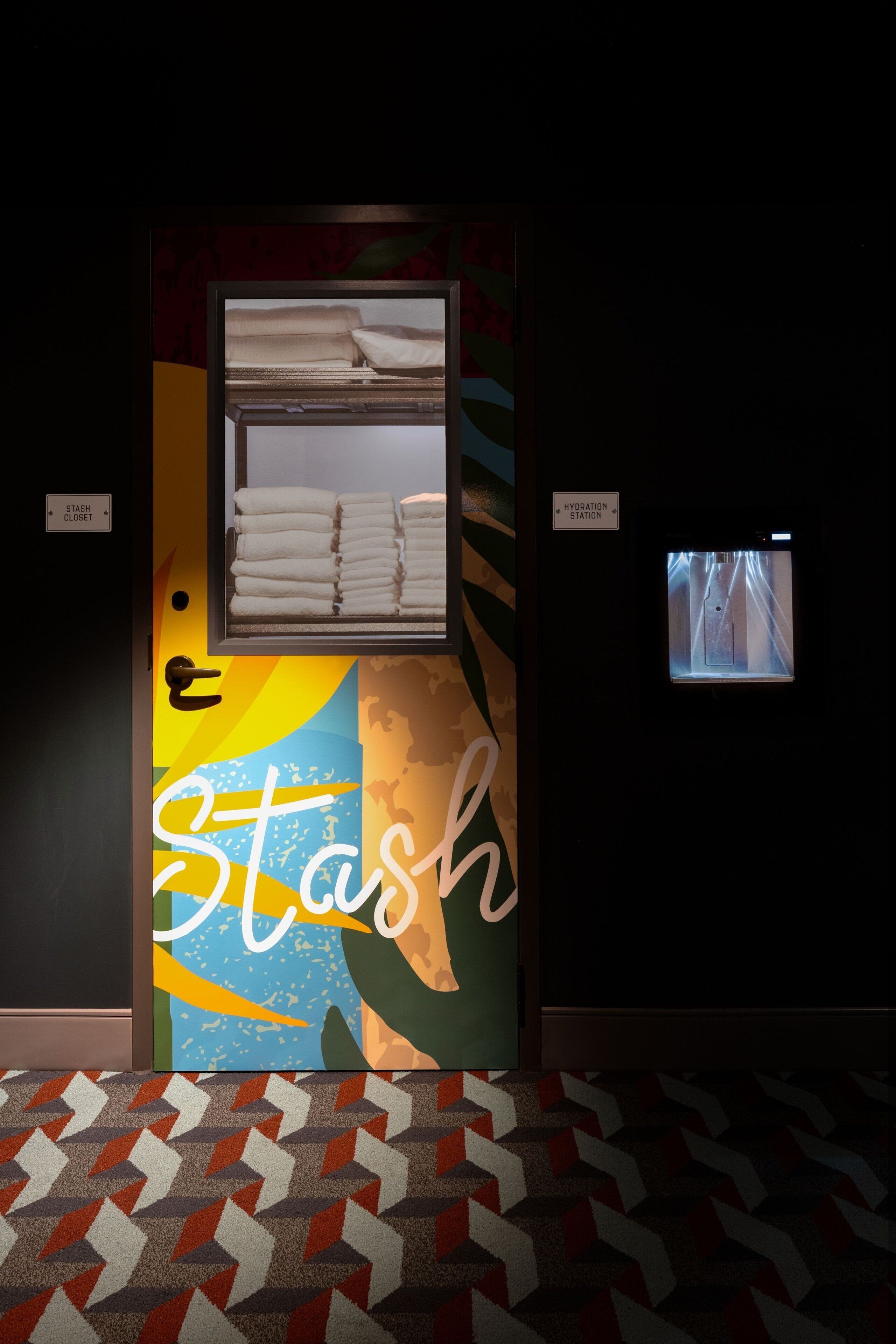 A hotel room with a bed, a window, and a wall-mounted TV. There is a sign indicating a stash closet and a hydration station.