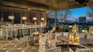 An outdoor bar area with wicker furniture, a bar counter stocked with bottles, and a city view at dusk.