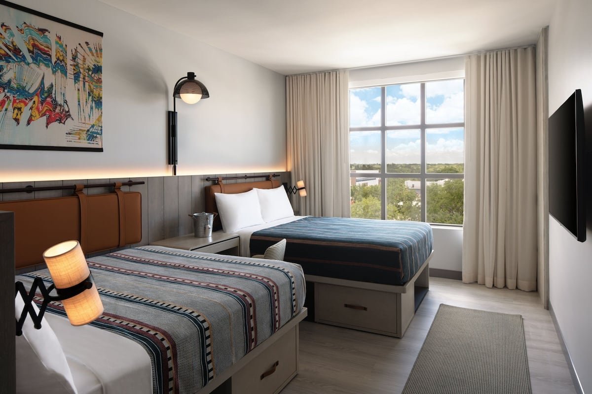 Ein Hotelzimmer mit zwei Betten, einem großen Fenster mit Vorhängen, einem an der Wand befestigten Fernseher und einer Wandleuchte.
