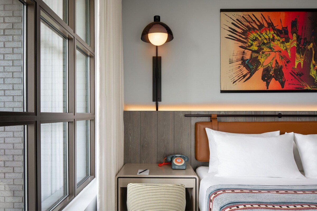 Une chambre d’hôtel dotée d’une grande fenêtre, d’une lampe de chevet, d’un téléphone, d’un lit avec du linge de lit blanc et d’un tableau abstrait aux couleurs vives accroché au mur.
