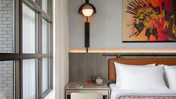 Ein Hotelzimmer mit großem Fenster, einer Nachttischlampe, einem Telefon, einem Bett mit weißen Leinentüchern und einem farbenfrohen abstrakten Gemälde an der Wand.