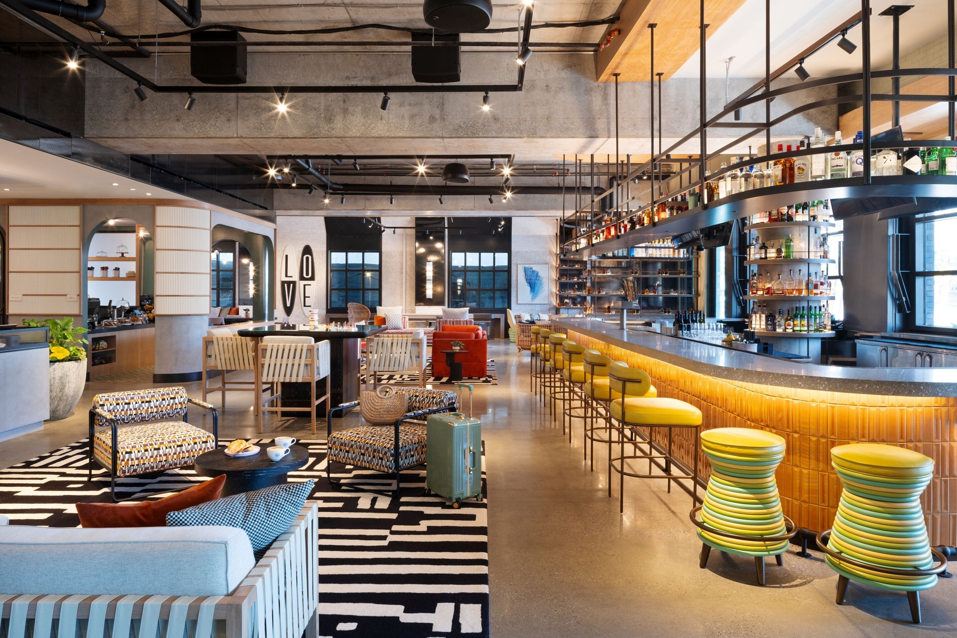 A modern bar area with a bar counter, stools, and a variety of bottles. There are seating areas with sofas and chairs, and a large window providing natural light.