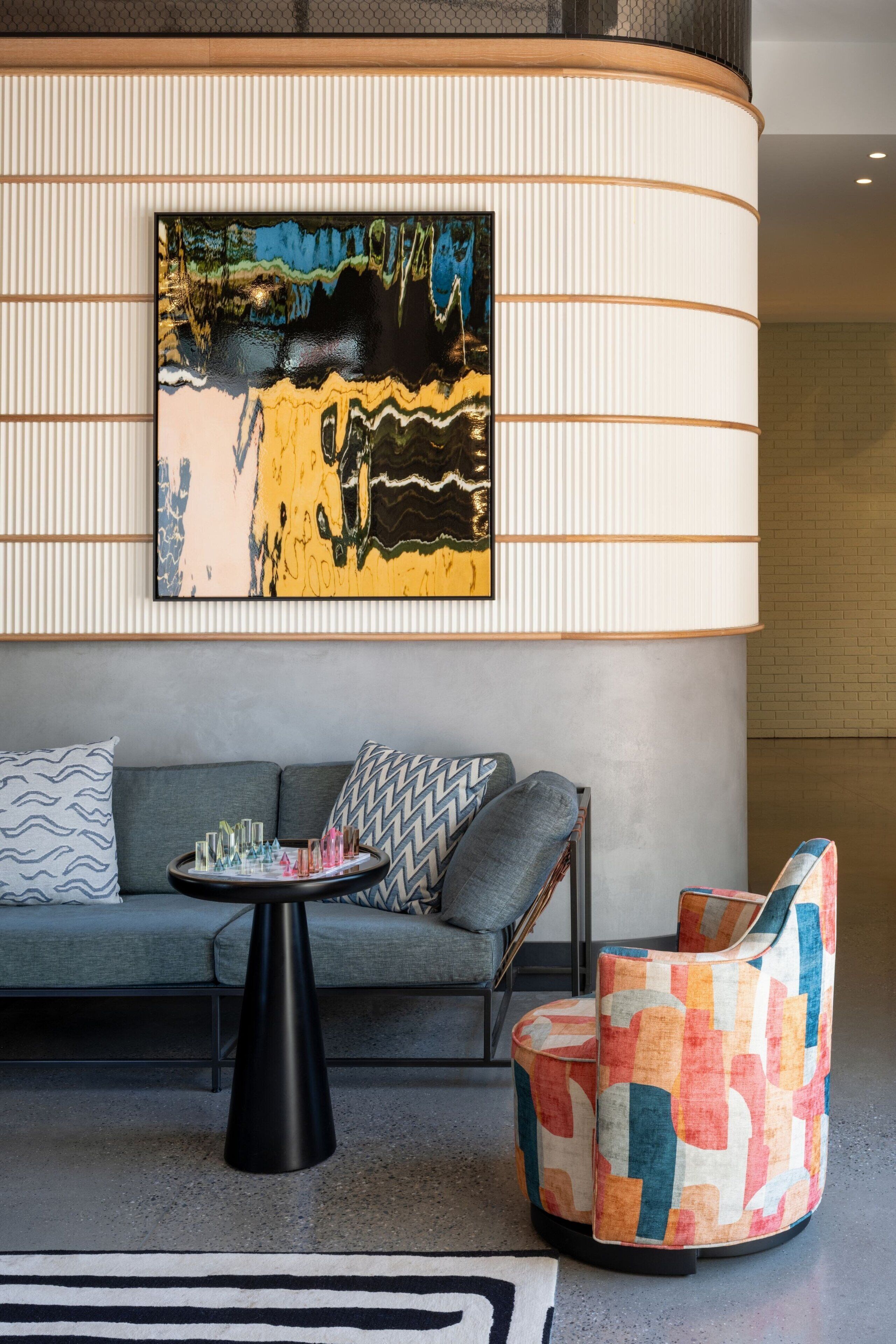A modern living room with a grey sofa, patterned cushions, a black coffee table, and a colorful armchair. A large abstract painting is displayed on the wall above the sofa.
