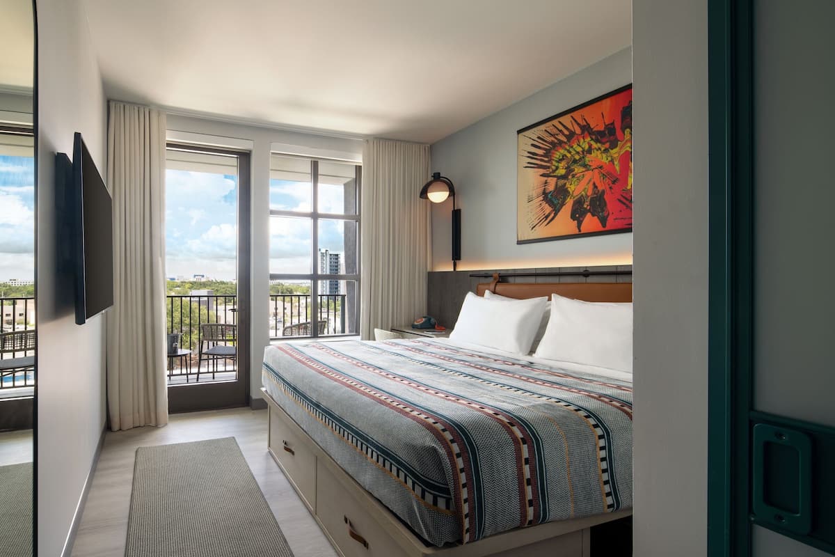 Ein Hotelzimmer mit einem großen Bett, einem Flachbildfernseher, einem Balkon mit Blick auf die Stadt und einem Gemälde an der Wand.