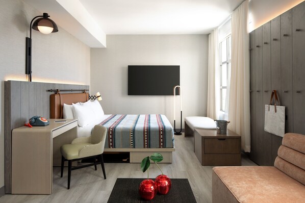A modern hotel room with a bed, desk, chair, bedside table, and a TV mounted on the wall.