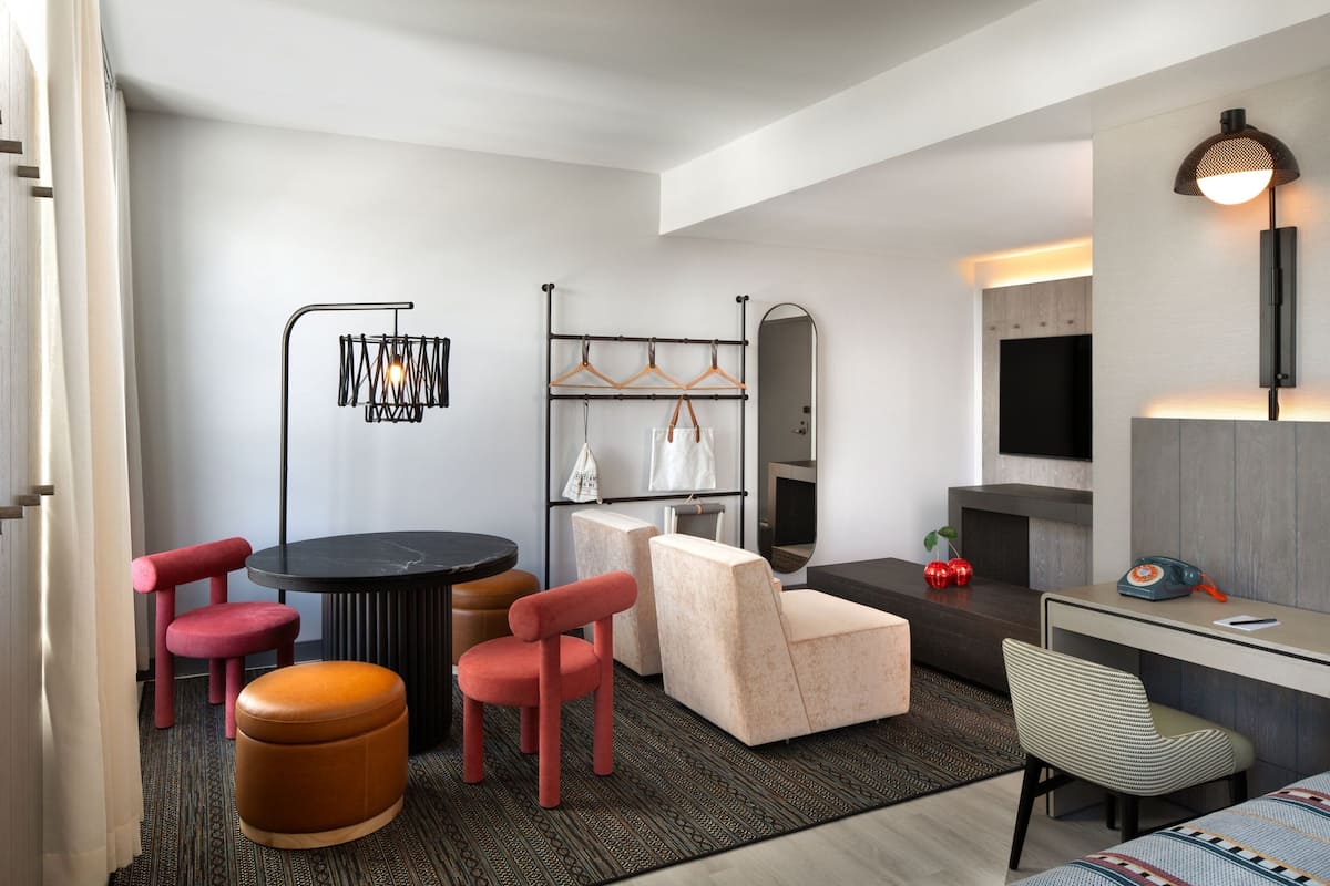 Ein modernes Hotelzimmer mit einem runden Esstisch, roten Stühlen, einer Couch, einem kleinen Hocker, einem Spiegel, einem Fernseher, einem Schreibtisch mit Lampe und einem Kleiderhaken.