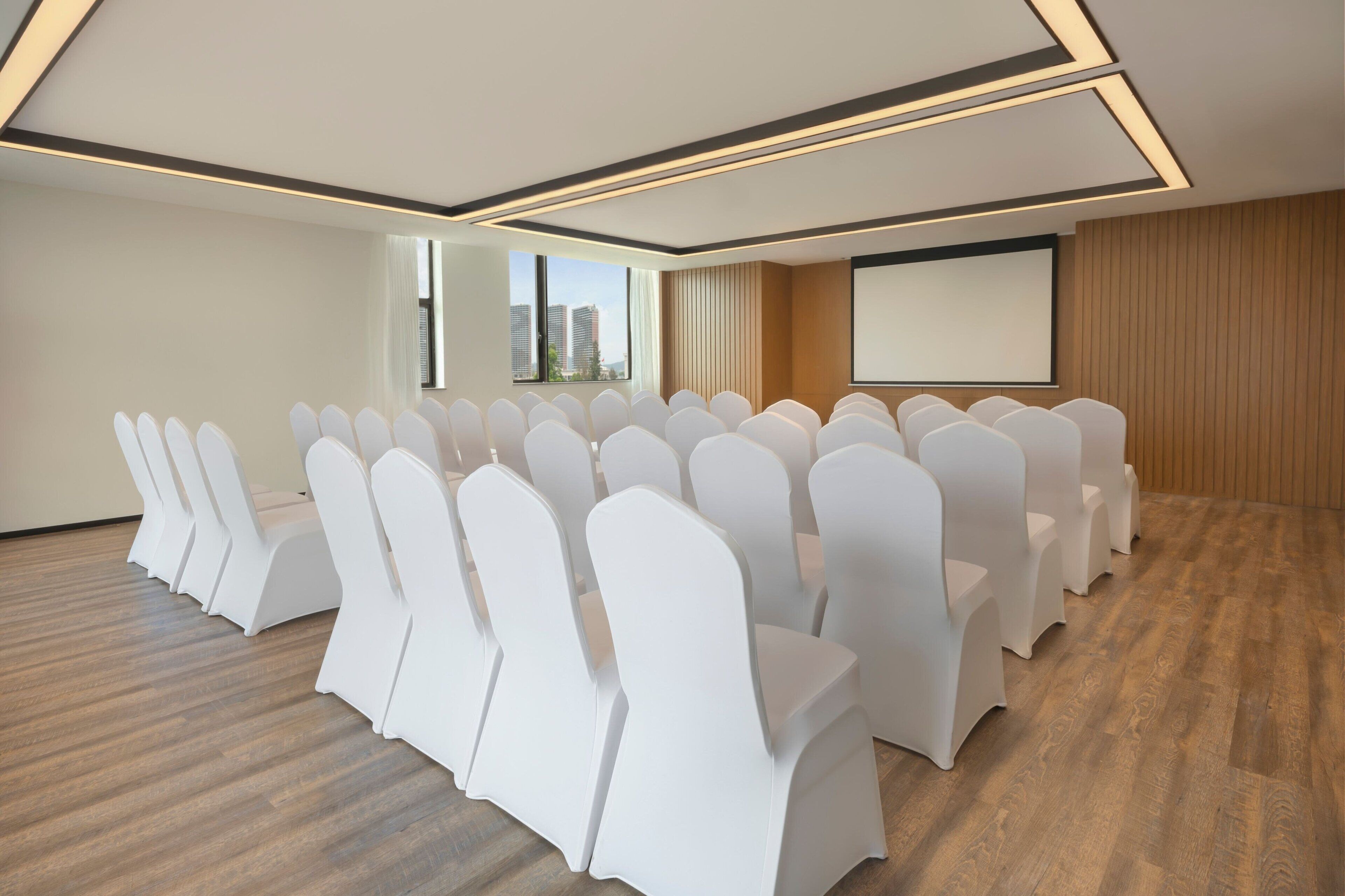 A conference room with rows of white chairs, a projector screen, and a large window with a city view.