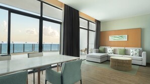 Une chambre d’hôtel moderne dotée d’une grande fenêtre donnant sur la mer, d’un coin repas avec une table et des chaises, d’un ensemble de canapés et d’une table basse ronde.
