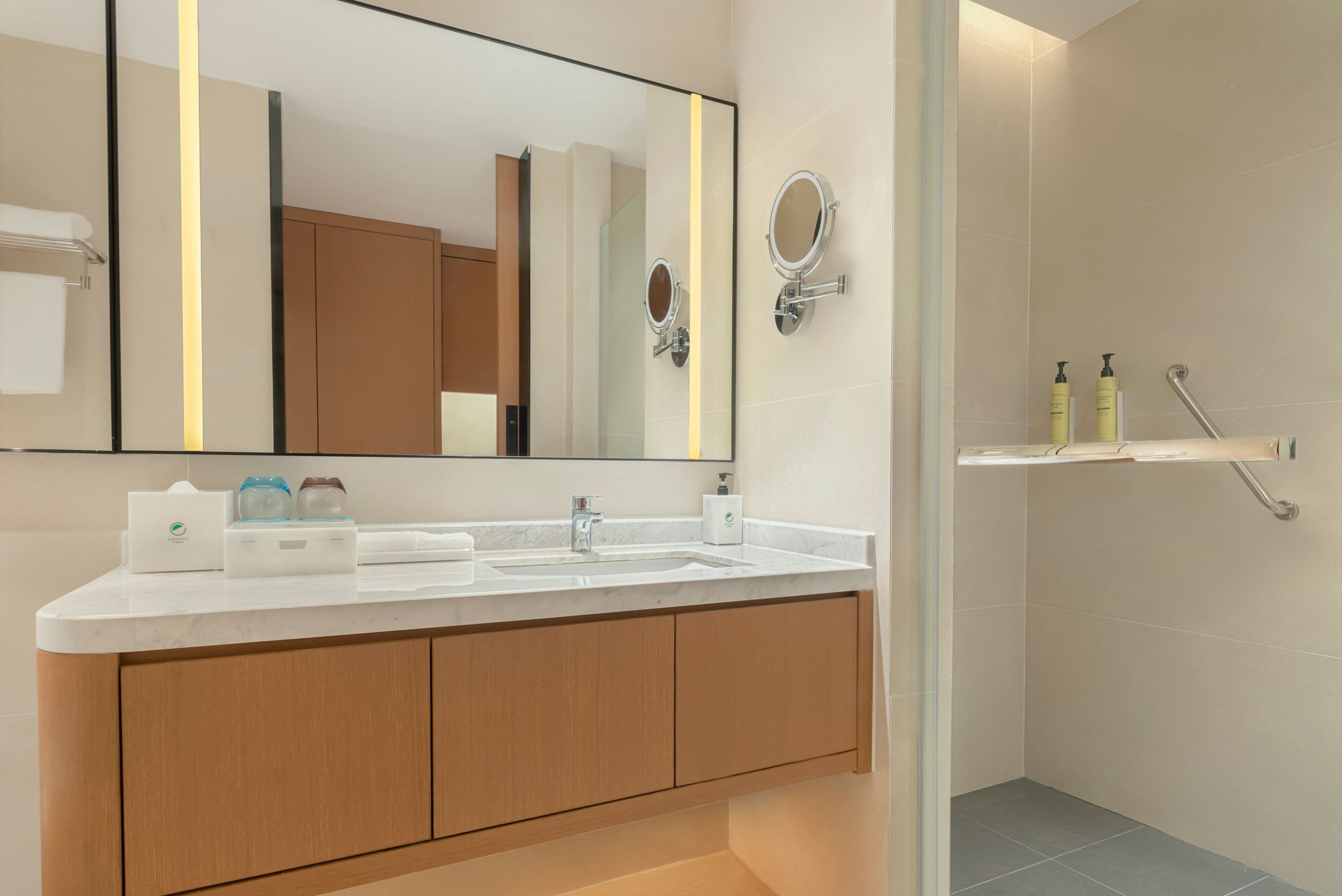 A modern bathroom with a large mirror, dual sinks, wooden cabinets, and a shower area.
