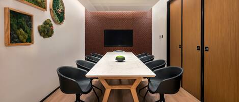 Uma sala de conferência com uma mesa comprida, cadeiras pretas, uma televisão na parede e uma porta de madeira.