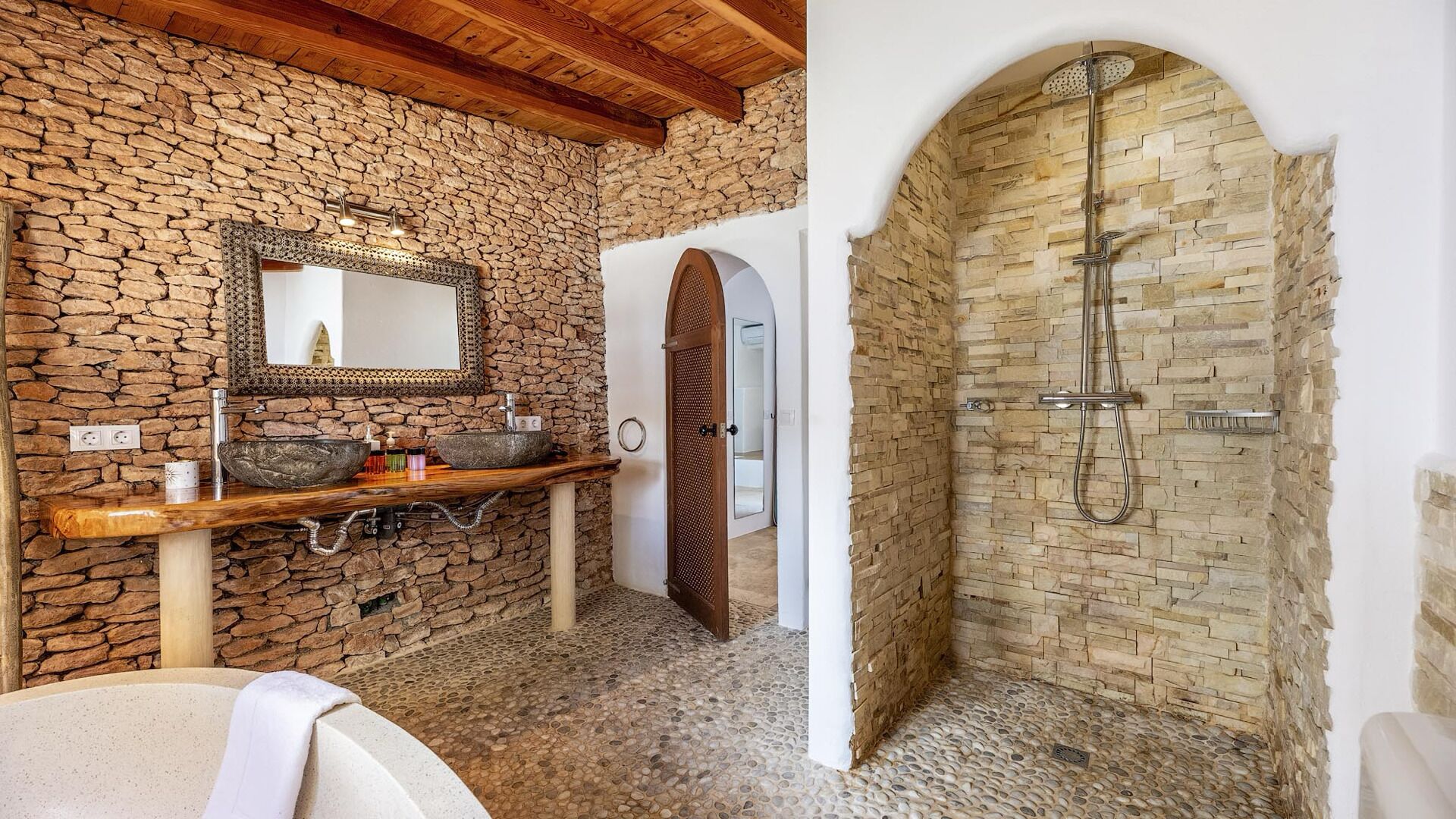 A bathroom with a stone wall, a wooden vanity, a large mirror, and a shower area with a stone wall.