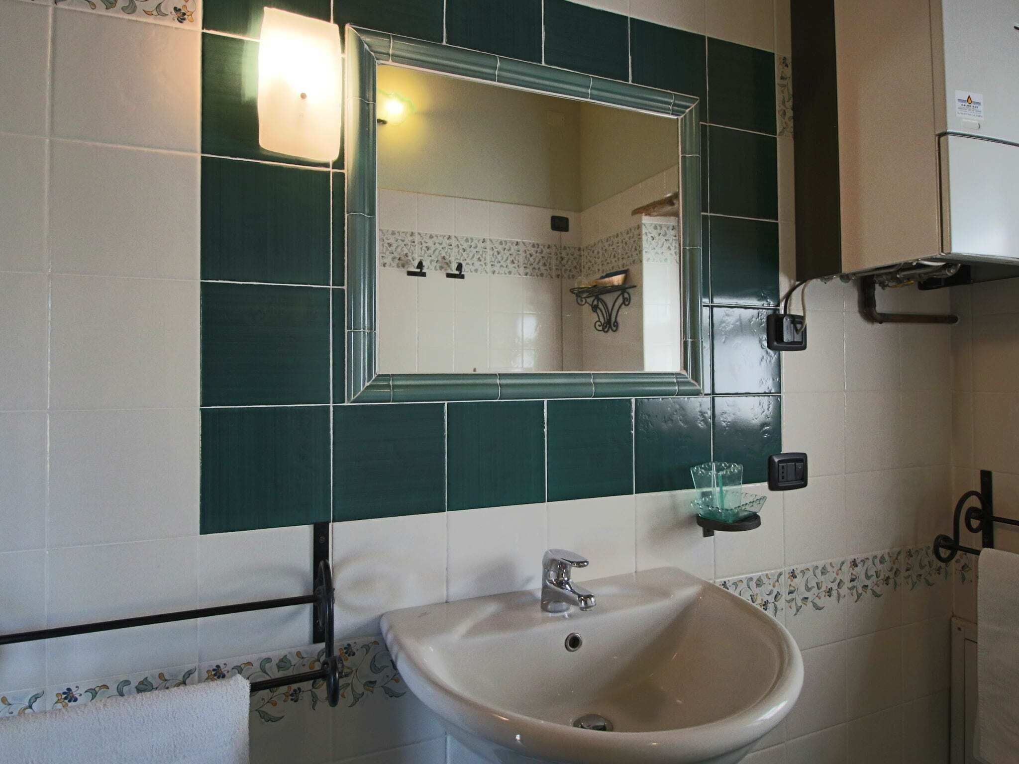 Ein Badezimmer mit Waschbecken, grünen Fliesen, einem Spiegel und einer kleinen Ablage mit einem Glasbehälter.